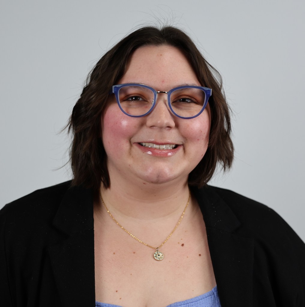 A brunette nonbinary person named Corian Zacher smiling and wearing blue round glasses and a sun necklace.